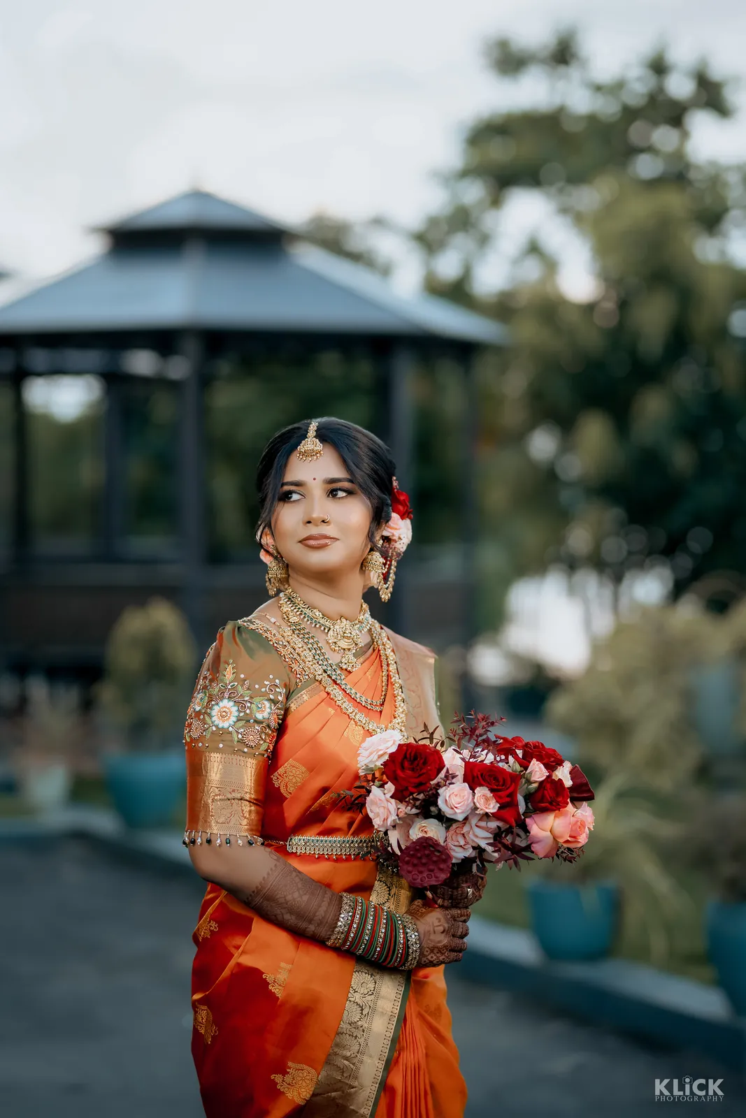 Bride in a red saree holding a bouquet beside a garden pavilion