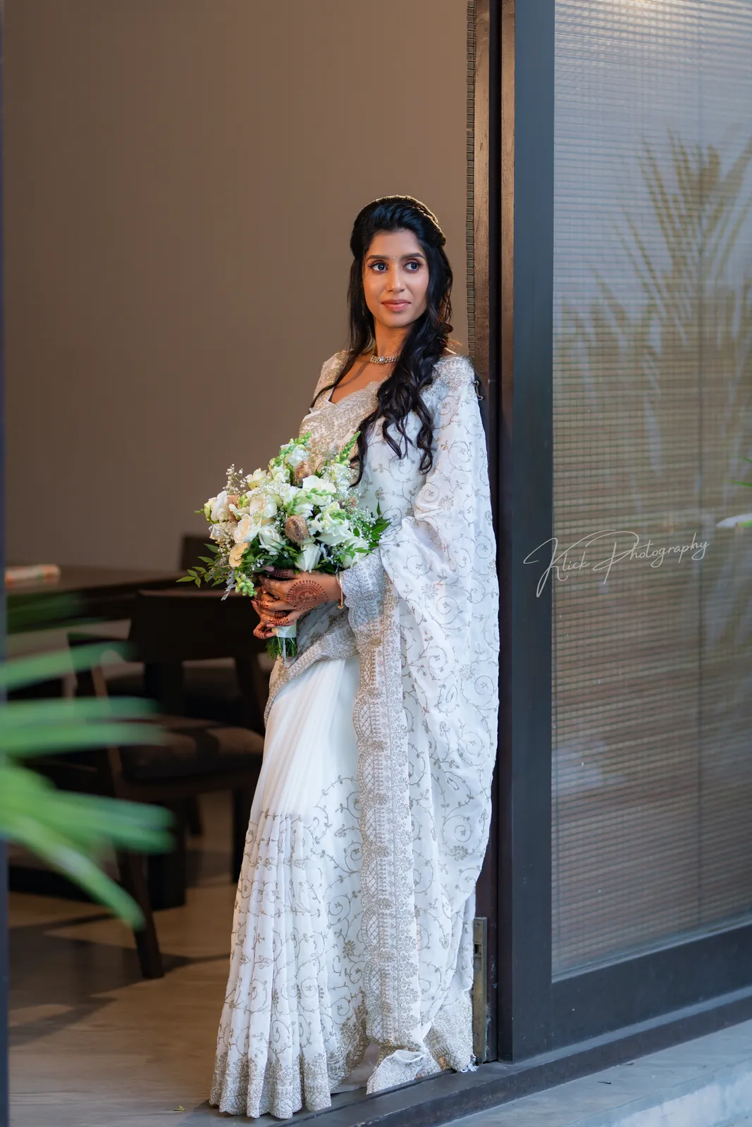 Bride in a white saree holding a bouquet by a window
